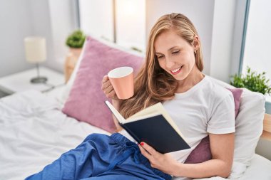 Genç sarışın kadın yatak odasında bir fincan kahve içip kitap okuyor.