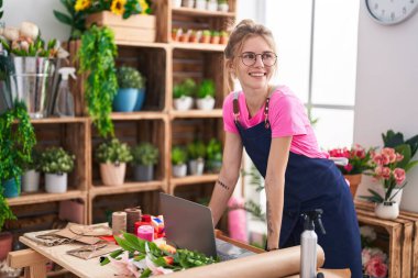 Çiçekçide dizüstü bilgisayar kullanarak gülümseyen genç sarışın kadın.