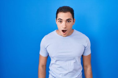 Young hispanic man standing over blue background afraid and shocked with surprise and amazed expression, fear and excited face. 