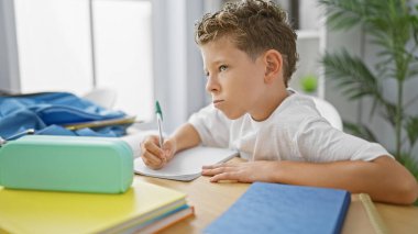 Sevimli sarışın öğrenci, öğrenmeye odaklanmış, kapalı sınıftaki masada oturmuş, akademik çalışmalara kafa yoruyor.