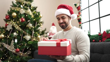 Evdeki Noel ağacının yanındaki koltukta oturan İspanyol genç adam hediyesini tutuyor.