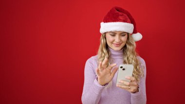 Yılbaşı şapkası takan genç sarışın kadın izole edilmiş kırmızı arka plan üzerinde akıllı telefon kullanıyor.