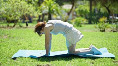 Orta yaşlı bir kadın parkta yoga minderinde geriniyor.