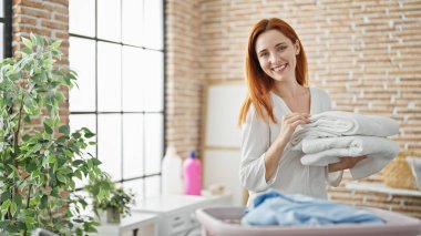 Çamaşırhanede katlanmış havluları tutan, kendine güvenen genç kızıl saçlı kadın.