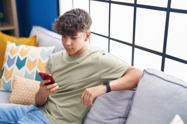 Evdeki kanepede oturan Latin kökenli genç akıllı telefon kullanıyor.