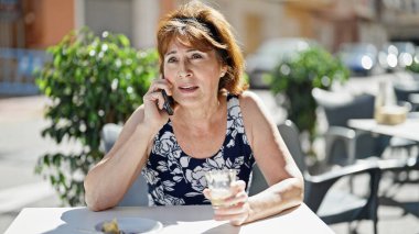 Kahve içen orta yaşlı bir kadın kahve dükkanının terasında akıllı telefondan konuşuyor.