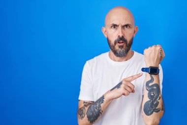 Mavi arka planda dikilen İspanyol dövmeli adam. Aceleyle saati, sabırsızlığı ve rahat bir ifadeyle kameraya bakıyor. 