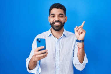 Sakallı İspanyol adam akıllı telefon kullanarak mesaj yazıyor. Yüzünde kocaman bir gülümseme var. Elinle kameraya bakıyor.. 