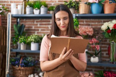 Young beautiful hispanic woman florist smiling confident reading notebook at flower shop