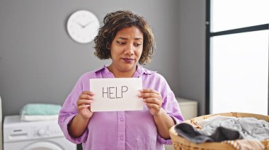 Genç, güzel, Latin bir kadın çamaşır odasında çamaşır ipine asılmış, üzgün bir ifadeyle yardım kağıdı tutuyor.