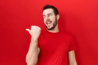 Gündelik kırmızı tişört giyen, gülümseyen, gülümseyen ve başparmağıyla yan tarafı gösteren İspanyol genç adam.. 