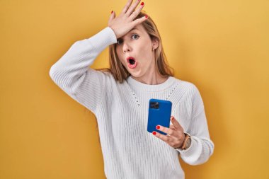 Young blonde woman using smartphone typing message surprised with hand on head for mistake, remember error. forgot, bad memory concept.  clipart