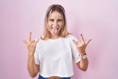 Pembe arka planda duran genç sarışın kadın çılgın bir ifadeyle el kaldırarak rock sembolü yapıyor. Müzik yıldızı. ağır müzik konsepti. 