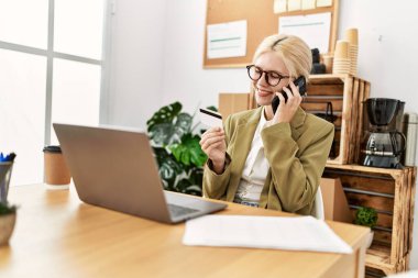 Genç sarışın iş kadını. Ofiste elinde kredi kartıyla akıllı telefondan konuşuyor.