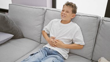 Sevimli sarışın çocuk mide ağrısı çekiyor, endişeli bir yüzle evde oturuyor, karnı ağrıyor.