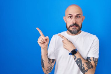 Mavi arka planda dikilen İspanyol dövmeli adam iki eliyle endişeli ve gergin, endişeli ve şaşırmış ifadesini işaret ediyor. 