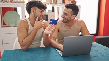 İki erkek alışveriş yapıyor. Laptop ve kredi kartıyla yemek odasında oturuyor.