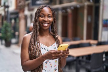 Afro-Amerikalı bir kadın kahve dükkanının terasında akıllı telefon kullanarak gülümsüyor.