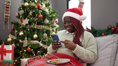 Saçları örgülü Afrikalı bir kadın akıllı telefon kullanıyor. Yılbaşı ağacının yanında oturuyor.