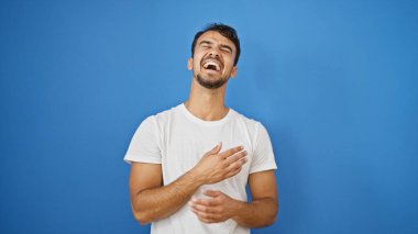 Genç İspanyol adam izole edilmiş mavi arka planda gülüp duruyor.