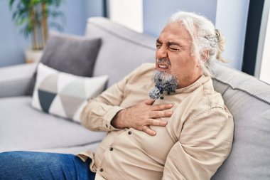 Middle age grey-haired man sitting on sofa suffering heart attack at home