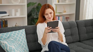 Young redhead woman using touchpad sitting on sofa at home