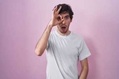 Pembe arka planda duran genç İspanyol adam şaşırmış yüz ifadesiyle şaşırmış, parmaklarının arasından bakan bir jest yapıyor. İnançsız ifade. 