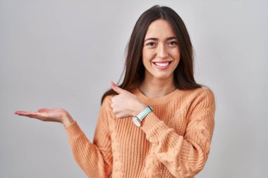 Beyaz arka plan üzerinde duran genç esmer kadın avuç içini gösteriyor ve baş parmaklarıyla iyi bir hareket yapıyor, mutlu ve neşeli gülümsüyor. 