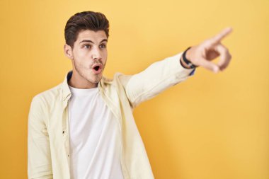 Young hispanic man standing over yellow background pointing with finger surprised ahead, open mouth amazed expression, something on the front 