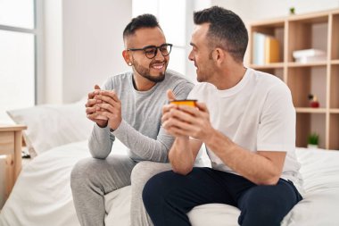 Two men couple drinking cup of coffee sitting on bed at bedroom