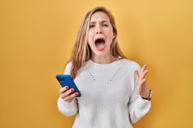 Genç sarışın kadın akıllı telefon kullanarak deli gibi mesaj yazıyor. Bağırıyor, bağırıyor, agresif ifadeler takınıyor ve kollar havada. hüsran kavramı. 