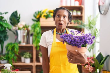 Çiçekçi dükkanında çalışan Afrikalı kadın elinde bitki tutarken ağzı açık kalmış, korkmuş ve hayrete düşmüş. 