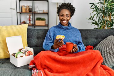 Afro-Amerikalı kadın kahvaltısını evde, kanepede yapıyor.