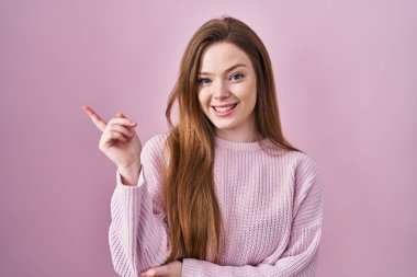 Young caucasian woman standing over pink background with a big smile on face, pointing with hand finger to the side looking at the camera. 