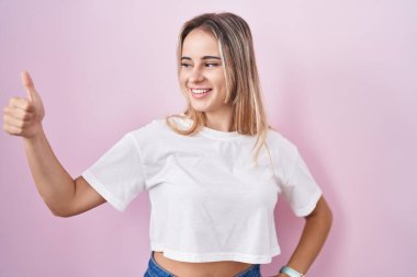 Pembe arka planda duran genç sarışın kadın. Gururlu görünüyor, gülümsüyor. Kenarda baş parmak hareketi yapıyor. 