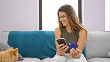 Kendinden emin, gülümseyen İspanyol bir kadın evde online alışveriş yapıyor oturma odasındaki kanepede köpeğiyle oturuyor, akıllı telefon ve kredi kartı kullanıyor..