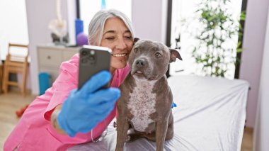 Gülümseyen orta yaşlı veteriner veteriner veteriner veteriner veteriner veteriner kliniğinde kıdemli bir köpekle selfie çekiyor.