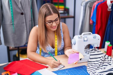 Young beautiful hispanic woman tailor writing on notebook holding cloth at tailor shop
