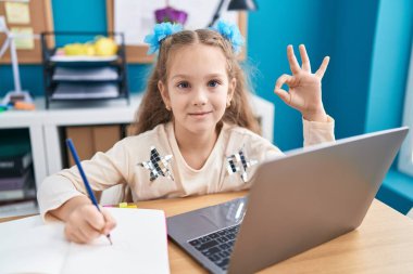 Masada oturmuş laptopla ödev yapan küçük bir kız parmaklarıyla imza atıp gülümsüyor ve mükemmel bir sembol oluşturuyor. 