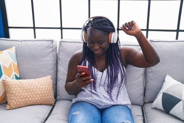 African american woman dancing and listening to music sitting on sofa at home