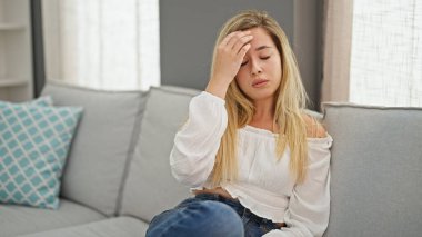 Evdeki kanepede oturan genç sarışın kadın baş ağrısı çekiyor.