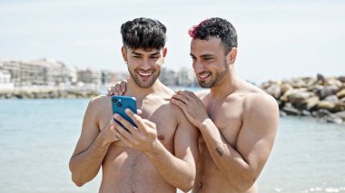 İki erkek turist kumsalda akıllı telefon kullanarak gülümsüyor.