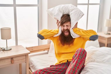 Young redhead man covering ears for noise at bedroom