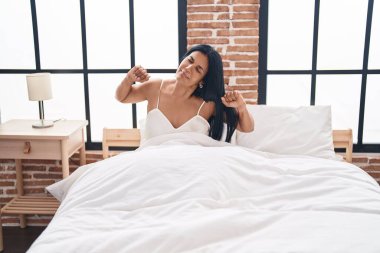 Middle age hispanic woman waking up stretching arms at bedroom