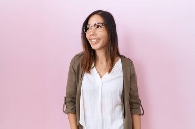 Orta yaşlı Çinli kadın pembe arka planda gözlük takıyor. Yüzünde gülümseme, doğal bir ifade var. Kendinden emin gülen. 