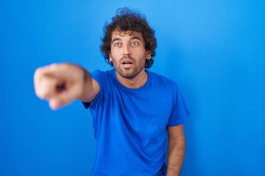 Latin kökenli genç bir adam mavi arka planda dikiliyor. Parmağı önde, ağzı açık, şaşırmış bir ifade. Ön tarafta bir şey var. 