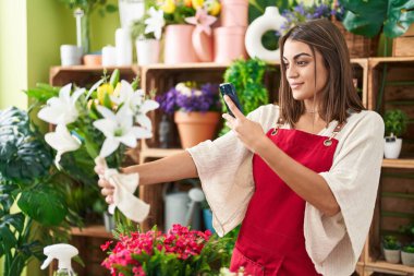 Young beautiful hispanic woman florist make photo to flowers by smartphone at flower shop