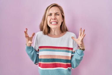 Young blonde woman standing over pink background crazy and mad shouting and yelling with aggressive expression and arms raised. frustration concept. 