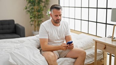 Beyaz saçlı, akıllı telefon kullanan, yatakta oturan bir adam.