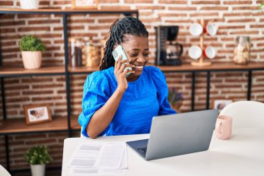 African american woman using laptop talking on smartphone at home